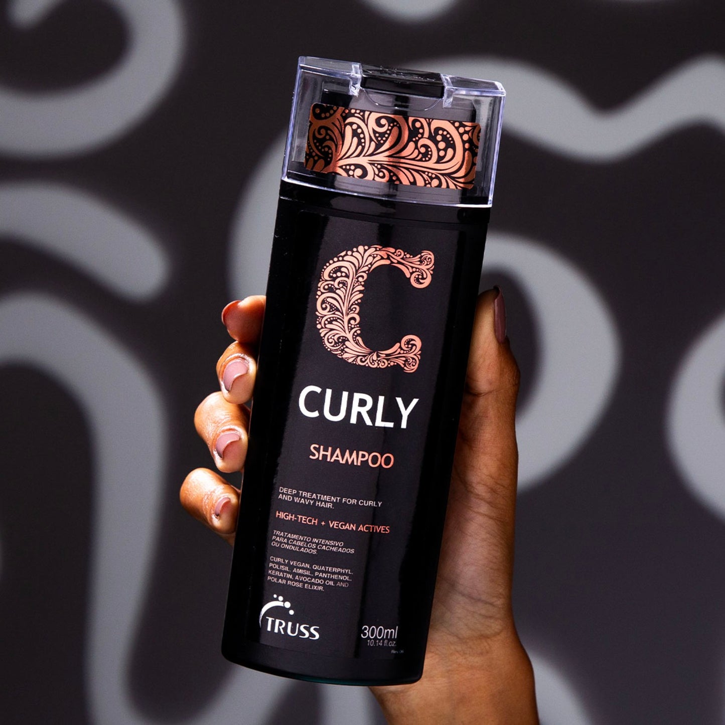 A imagem mostra uma mão segurando um frasco de shampoo da marca Truss, da linha Curly. O frasco é preto e tem uma tampa transparente, com 300ml.